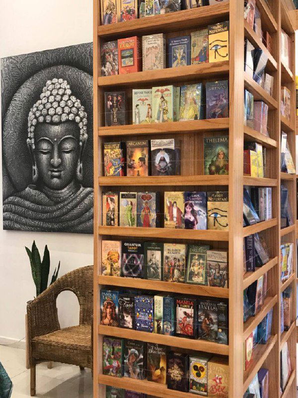 Угловой стеллаж с множеством колод Таро на фоне декора: изображение Будды и плетёное кресло — сочетание магазинной витрины и уютной выставки.