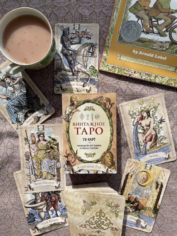 Коробка и карты колоды «Винтажное Таро» среди разбросанных карт и чашки с напитком: вид сверху показывает дизайн карт и атмосферу домашней практики Таро.
