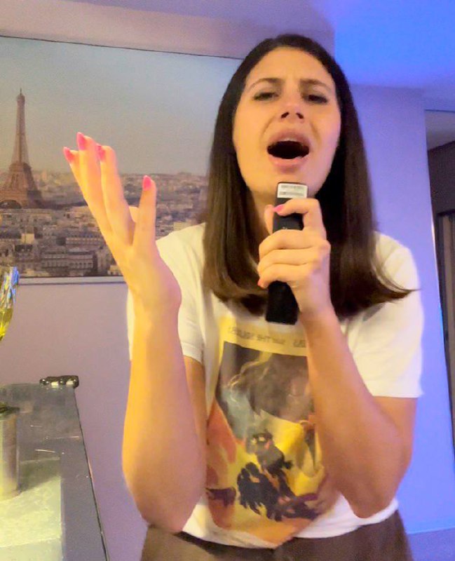 Женщина поёт эмоционально, широко раскрывает рот, держит микрофон в руках; на стене позади видна картина с Эйфелевой башней.