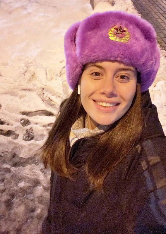 Селфи женщины в фиолетовой шапке-ушанке на снежной улице; улыбающееся лицо на фоне зимнего города, живой и личный кадр.