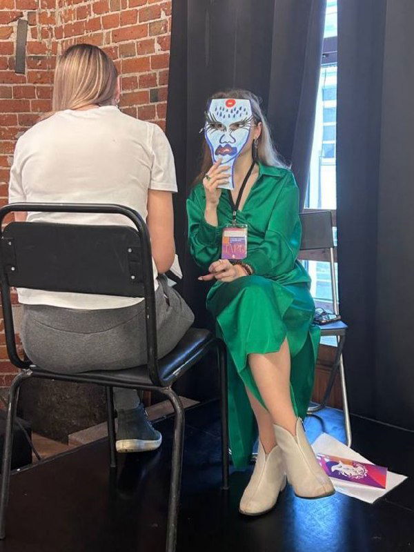 Участница в зелёном платье сидит на стуле и держит маску перед лицом, рядом кресло собеседника и материалы тренинга в комнате с кирпичными стенами.