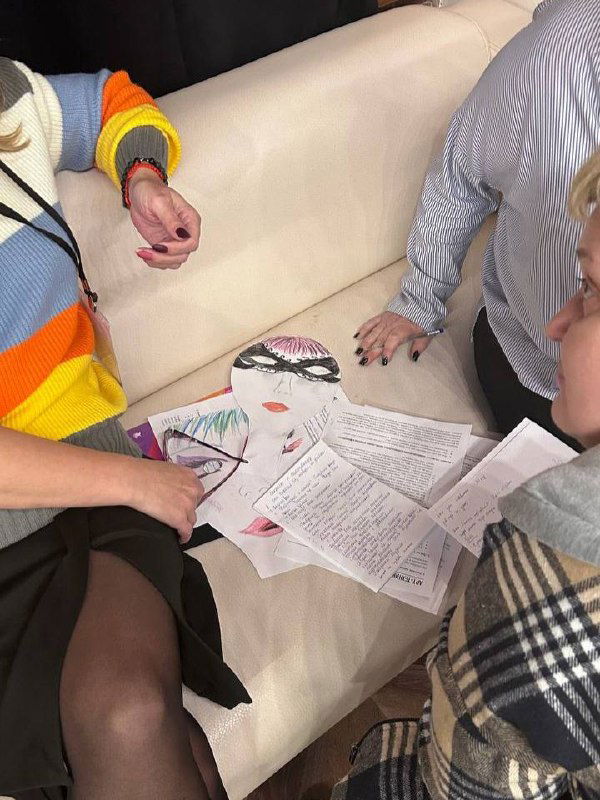 Группа людей около дивана рассматривает листы и маски, на коленях видны заметки и рисунки — коллективная работа в ходе занятия.