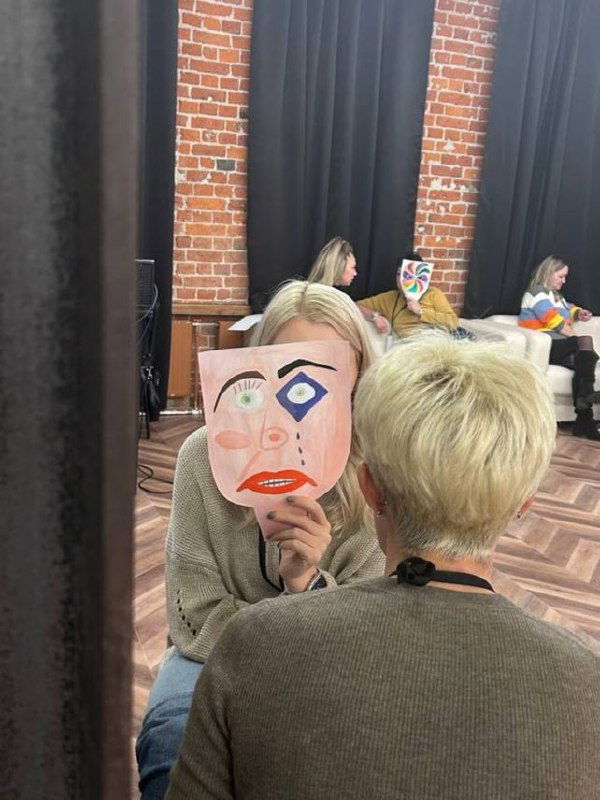 Две участницы сидят лицом друг к другу, одна держит нарисованную маску перед лицом — психологическое упражнение на взаимодействие.