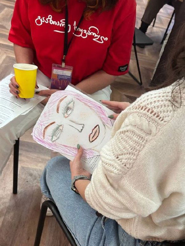 Крупный план рук участницы, держащей карандаш и бумажную маску с начерченными глазами и чертами лица — процесс создания маски.