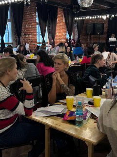 Широкий план зала с участниками, сидящими за столами в группах; видно оживлённую дискуссию и материалы тренинга на столах.