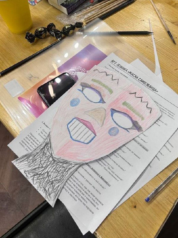 На столе лежит бумажная маска с раскрашенным лицом поверх листа с заданиями и брошюрой тренинга — рабочий набор участника.