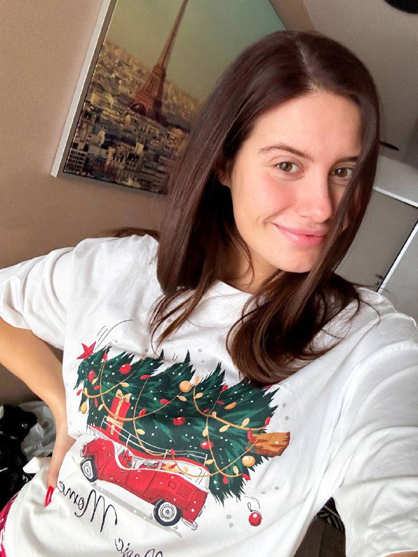 Крупный план женщины в той же белой футболке с ёлкой, естественная улыбка на лице, на фоне интерьерной картины с изображением Парижа.
