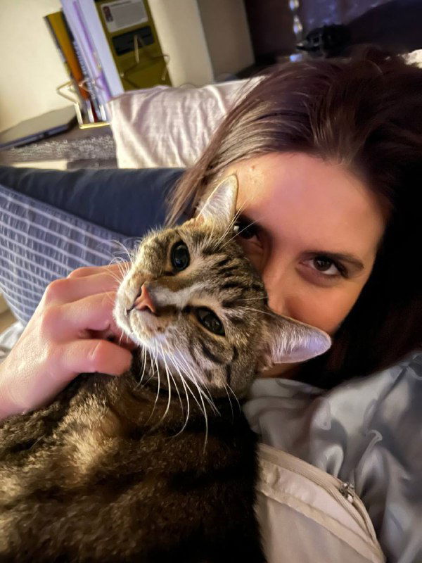 Крупный план: женщина держит и прижимает к лицу полосатого кота, видны глаза животного и элементы спальни на фоне.