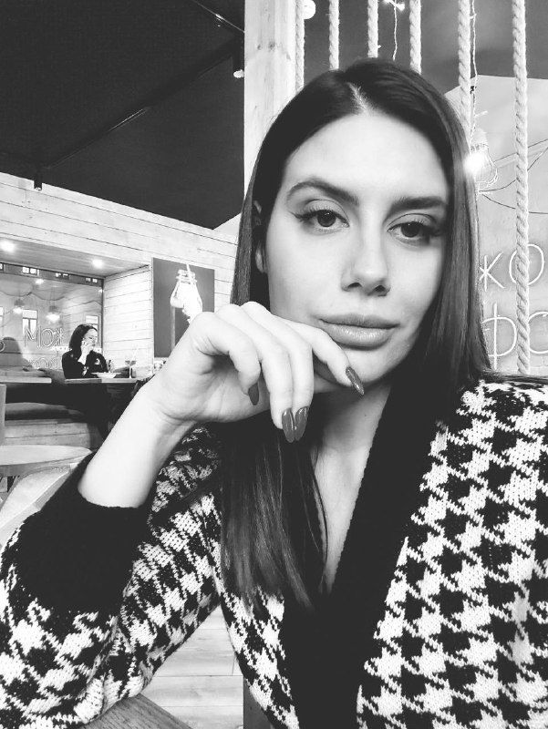 Чёрно‑белый фотопортрет девушки в кофейне, рука у подбородка, держащая взгляд в камеру; стильный узор одежды заметен на снимке.