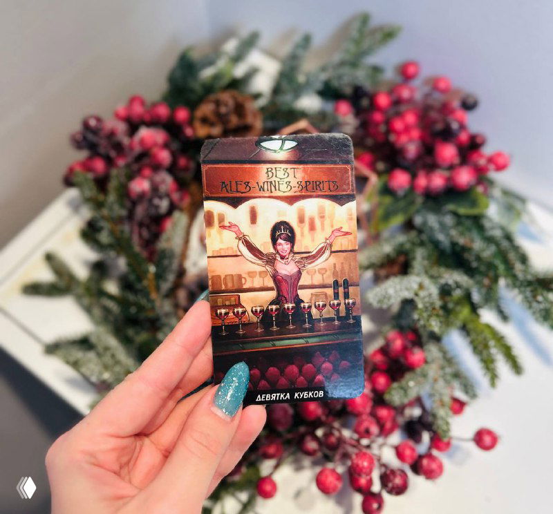 Рука держит другую карту Таро с изображением сцены в интерьере на фоне еловых веток и красных ягод; крупный план.