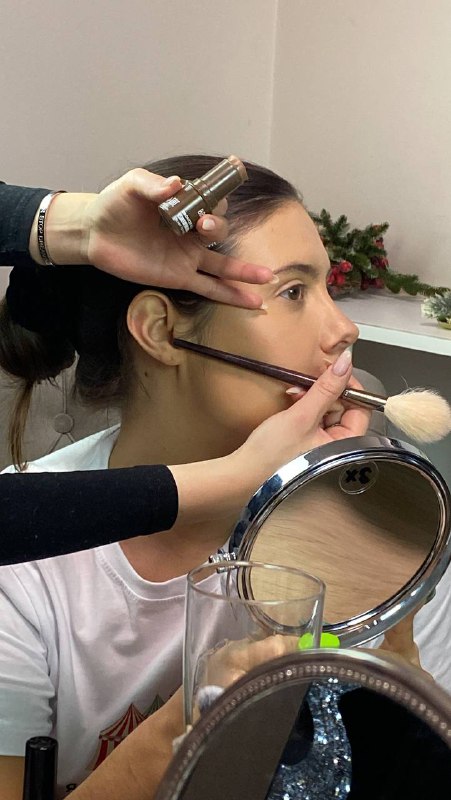 Крупный план процесса: визажист наносит кистью макияж на лицо модели, видны руки мастера, зеркала и косметические принадлежности в студии с мягким светом.