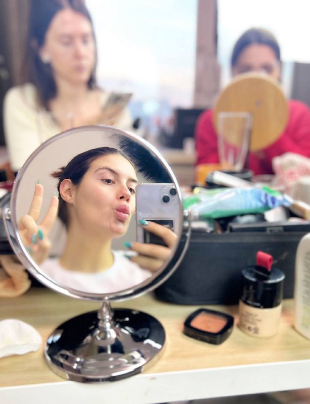 Отражение в настольном зеркале: девушка делает селфи на фоне косметики и мастериц, кадр передаёт живую атмосферу девичника и подготовки к ролику для YouTube.