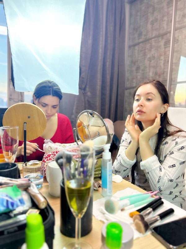 Две подруги у зеркала примеряют образы: на столе напиток, бьюти-продукты и аксессуары — уютный интерьер студии и совместная практика макияжа.