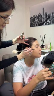 Портретный кадр работы визажиста: мастер растушёвывает тени и наносит румяна клиентке, видно зеркало, инструменты и професcиональный подход в студийной обстановке.