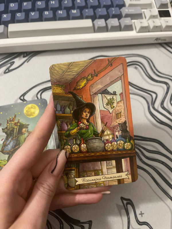 Рука держит карту Восьмерка Пентаклей из колоды Таро Ежедневной Ведьмы; видна поверхность стола и элементы рабочего места.