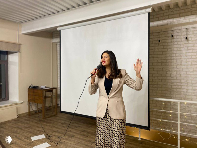 Ведущая с микрофоном на фоне экрана, она объясняет методику: профессиональная подача материала в пространстве для лекций и практик.