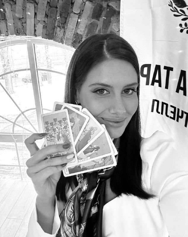 Чёрно-белое фото женщины с раскрытой колодой Таро на фоне кирпичной арки и баннера; контраст подчёркивает карты и лицо.