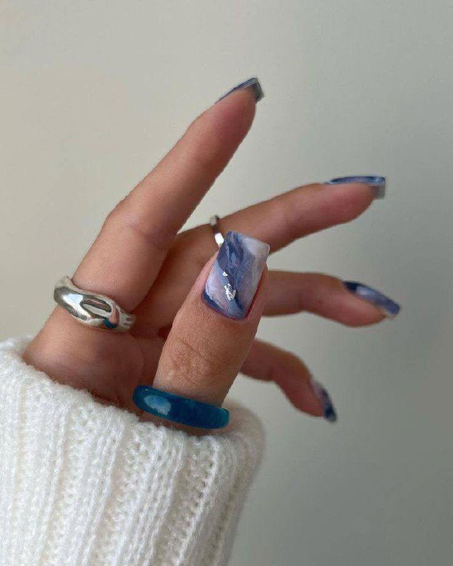 Рука с мраморным голубым дизайном на длинных квадратных ногтях, яркие синие акценты и массивные кольца, светлый нейтральный фон.
