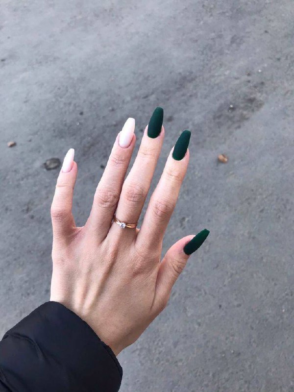 Рука с матовым тёмно-зелёным маникюром и пастельно-розовым акцентным ногтем; видно тонкое кольцо на пальце, уличный фон.