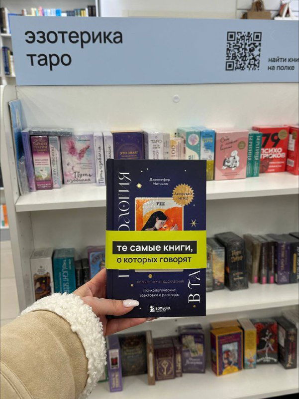 Фото в книжном магазине: рука держит книгу автора на полке в разделе «эзотерика / таро», видны соседние издания и ценники.