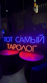 Неоновая вывеска «ТОТ САМЫЙ ТАРОЛОГ» в синих и розовых тонах, круглые столики и приглушённая обстановка зала — кадр с вечернего мероприятия в клубе.