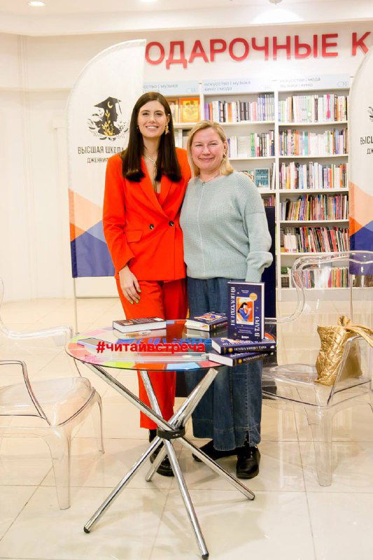 Автор в оранжевом костюме позирует с посетительницей рядом со столом презентации и баннером книжной школы на заднем фоне.