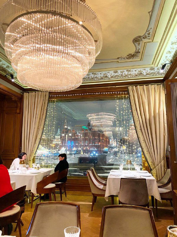 Интерьер ресторана с большой хрустальной люстрой и видом на иллюминированный городской пейзаж через панорамное окно; уютный зал.