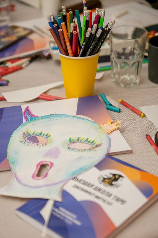 Рабочий стол тренинга: цветные карандаши в стакане, раскрашенная бумажная маска на листе и материалы по техникам.