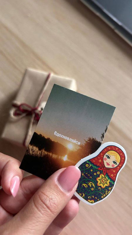 Рука держит маленькую иллюстрированную карточку и наклейку в виде матрёшки на фоне упакованной коробки — детали комплектности и эстетики колоды.