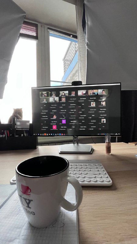 Фото рабочего места с монитором, где идёт онлайн‑тренинг: сетка участников на экране, кружка с напитком и клавиатура на столе вблизи.