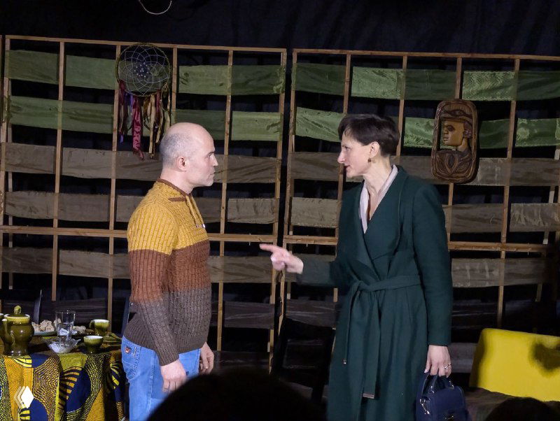 Двое актёров в диалоге на переднем плане на фоне деревянной клетки декорации; ощущение напряжения и сценического взаимодействия.