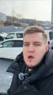 Мужчина делает селфи на фоне стоянки с припаркованными автомобилями; видны складские корпуса и зимняя улица.