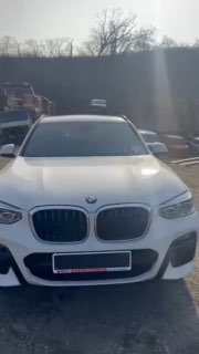 Фронтальный вид белого BMW X3 на открытой площадке; кадр из видеоотчёта с видимыми отражениями на капоте и городской средой на заднем плане.