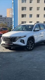 Вертикальный портретный кадр белого Hyundai Tucson 2020 спереди на парковке, отражения на кузове и городской фон, демонстрирующий общий внешний вид автомобиля перед отправкой в Иркутскую область.