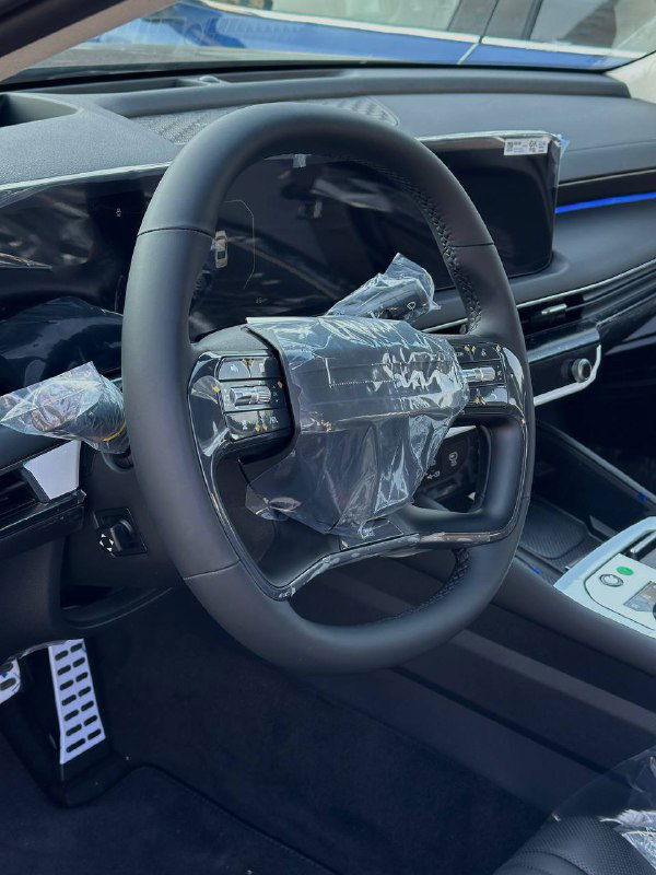 Рулевое колесо и часть приборной панели KIA K8 2024 в защитной плёнке: вид центральной зоны управления с элементами мультимедиа и кнопками на руле.