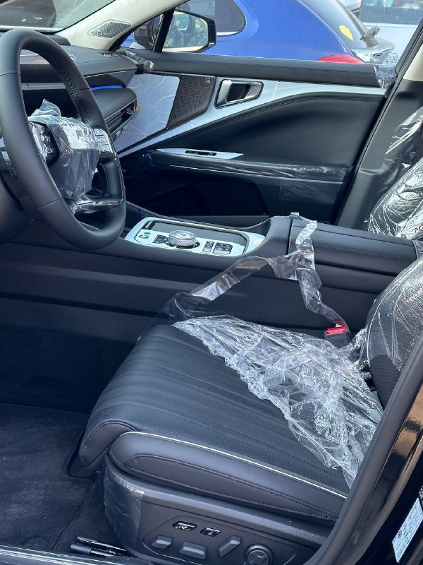 Пассажирская сторона салона KIA K8 2024: вид сиденья, дверной панели и элементов отделки в защитной плёнке, подчёркивающий внутреннюю отделку люксовой комплектации.