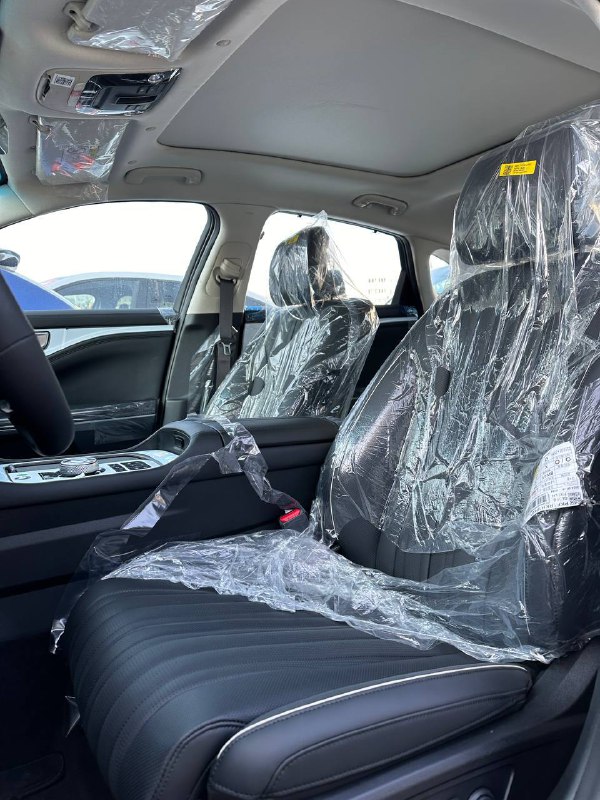 Фронтальные сиденья и потолок KIA K8 2024: снимок внутреннего пространства люксового седана с защитными плёнками на креслах и чистой отделкой.