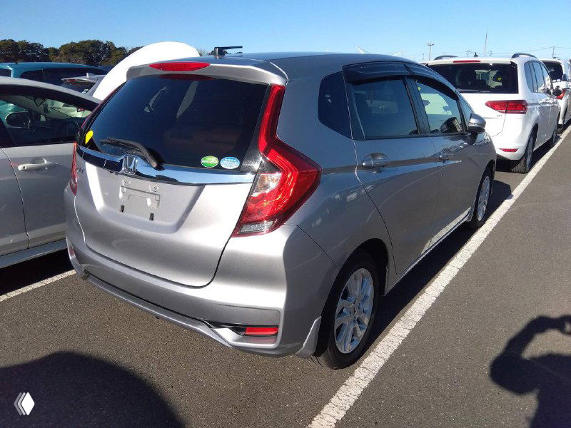 Задняя часть серебристого Honda Fit 2018 на парковке аукциона: вид сзади справа, открытый багажник отсутствует, задние фонари и форма кузова видны.