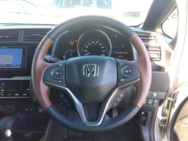 Крупный план руля Honda Fit 2018: кожаная отделка, элементы управления на руле, видно приборную панель и индикаторы на фоне.