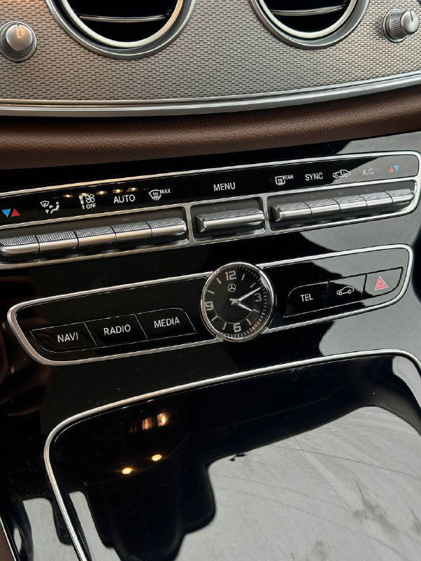 Крупный план центральной панели Mercedes E350: аналоговые часы, блок климата и кнопки мультимедиа в хромированных окантовках и чёрном глянце.