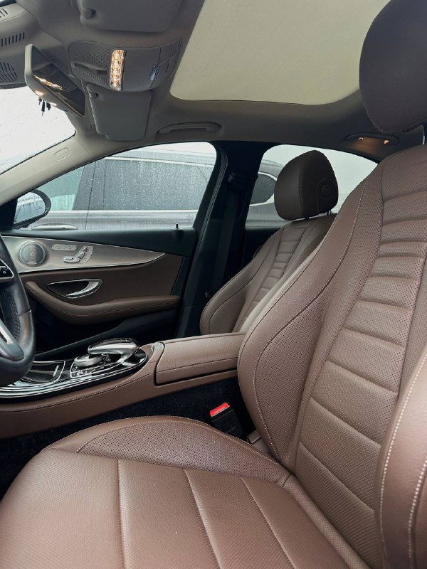 Передние сиденья Mercedes E350 в тёмно-коричневой коже с декоративной прострочкой и боковой поддержкой, вид со стороны двери на салон.