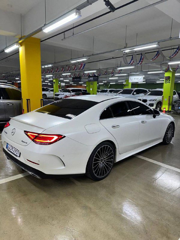 Вид сзади белого Mercedes‑Benz CLS450 2022 на крытой стоянке, подчёркнуты задние фонари, багажная часть и профиль кузова автомобиля, складской ракурс.
