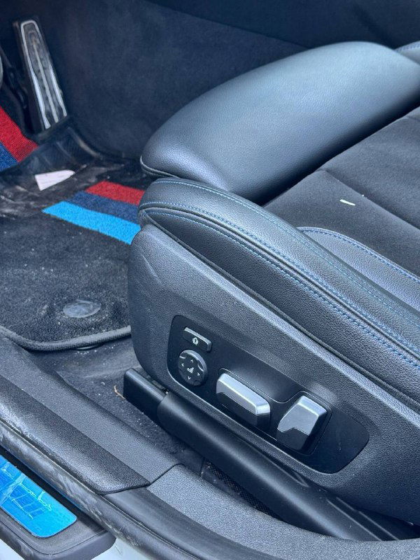 Кресло водителя BMW 320i M‑Sport — крупный план регулировок сиденья, боковой профиль отделки и фирменные цветовые акценты на коврике и обивке.