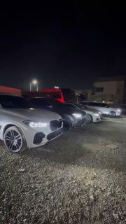 Ряд внедорожников и седанов на открытой стоянке ночью; кадр показывает автомобили, ожидающие отправки с крытой и открытой площадки в Южной Корее.