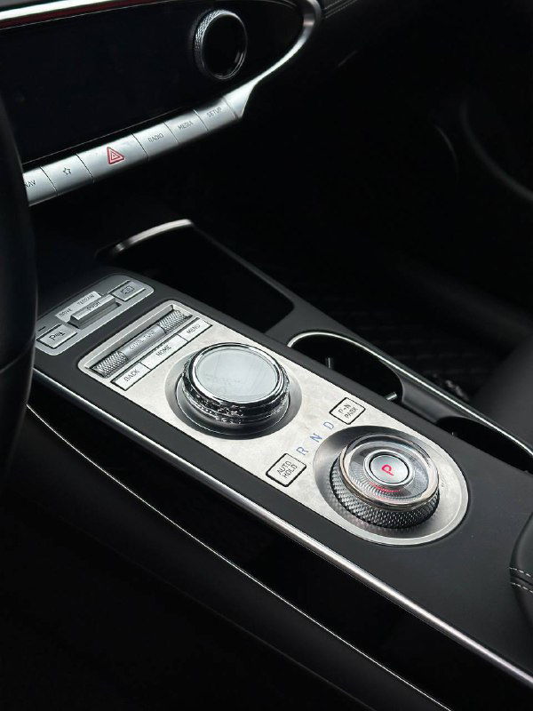 Крупный план центральной консоли Genesis GV70: селектор передач, поворотный контроллер и кнопки управления в салоне.