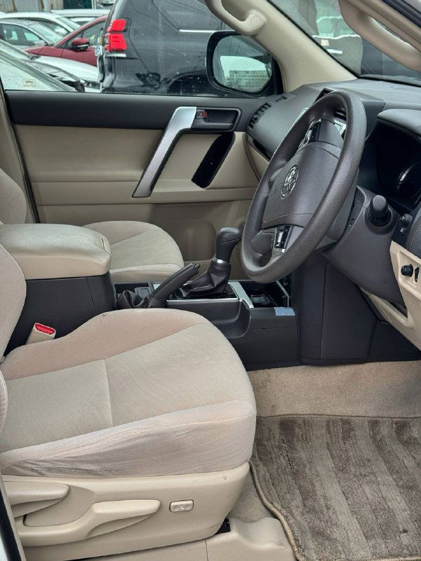 Интерьер водительского места Toyota Prado: рулевое колесо, панель приборов, переднее пассажирское кресло и центральный подлокотник в бежевой отделке