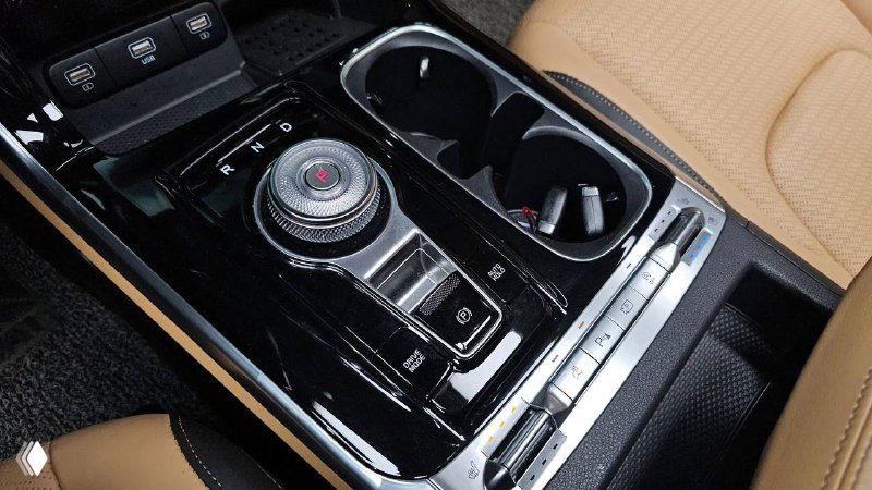 Центральная консоль KIA Carnival Signature 2021 с селектором передач, подстаканниками и отделкой в чёрном глянце на фоне бежевого салона.