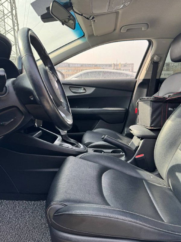 Вид на передние кресла и центральный тоннель KIA K3 с пассажирского ракурса, видна вместимость салона, подлокотник и элементы управления между сиденьями.