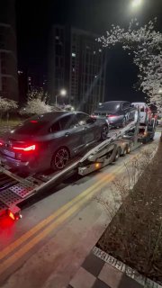 Вид сбоку на автопогрузку: на автовозе стоят Hyundai, BMW и другие машины, ночная улица с фонарями и цветущими деревьями в Корее.