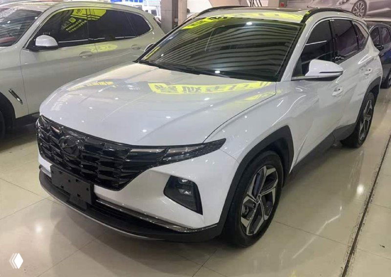 Крупный план передней части белого Tucson в автосалоне, чистый бампер и кузов без номерного знака, подчёркнутые линии фар и дневных ходовых огней.